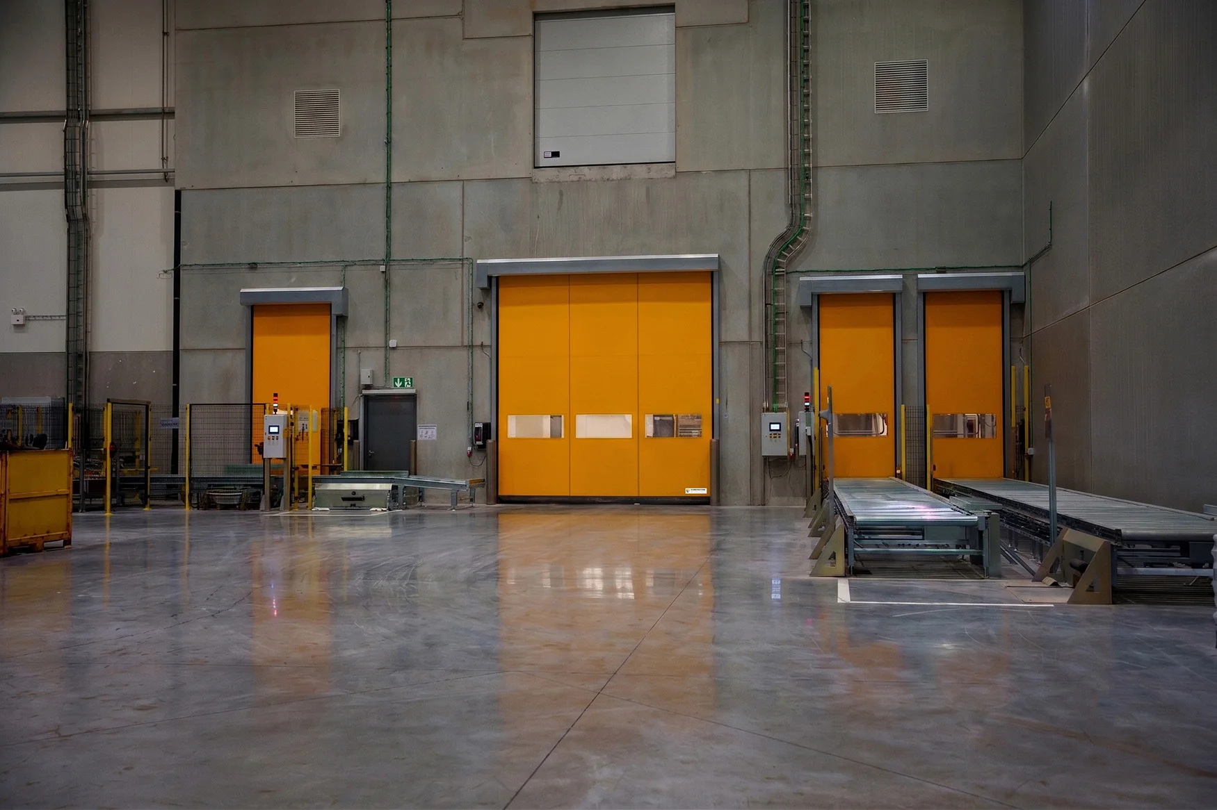 Industrieel magazijn met betonnen vloer en grijze wanden met drie oranje laaddeuren en gele stapelaar links.