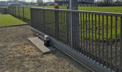 Grijze metalen toegangspoort met verticale spijlen op een betonnen fundering in een weiland met groene grasvelden.