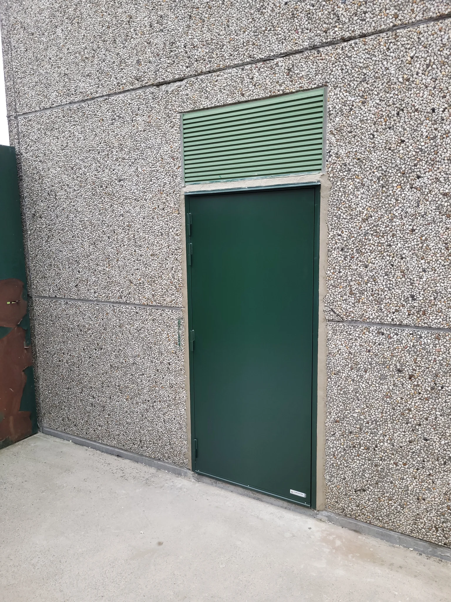 Groene branddeur met ventilatierooster in grijze steenachtige gevel van een modern gebouw.
