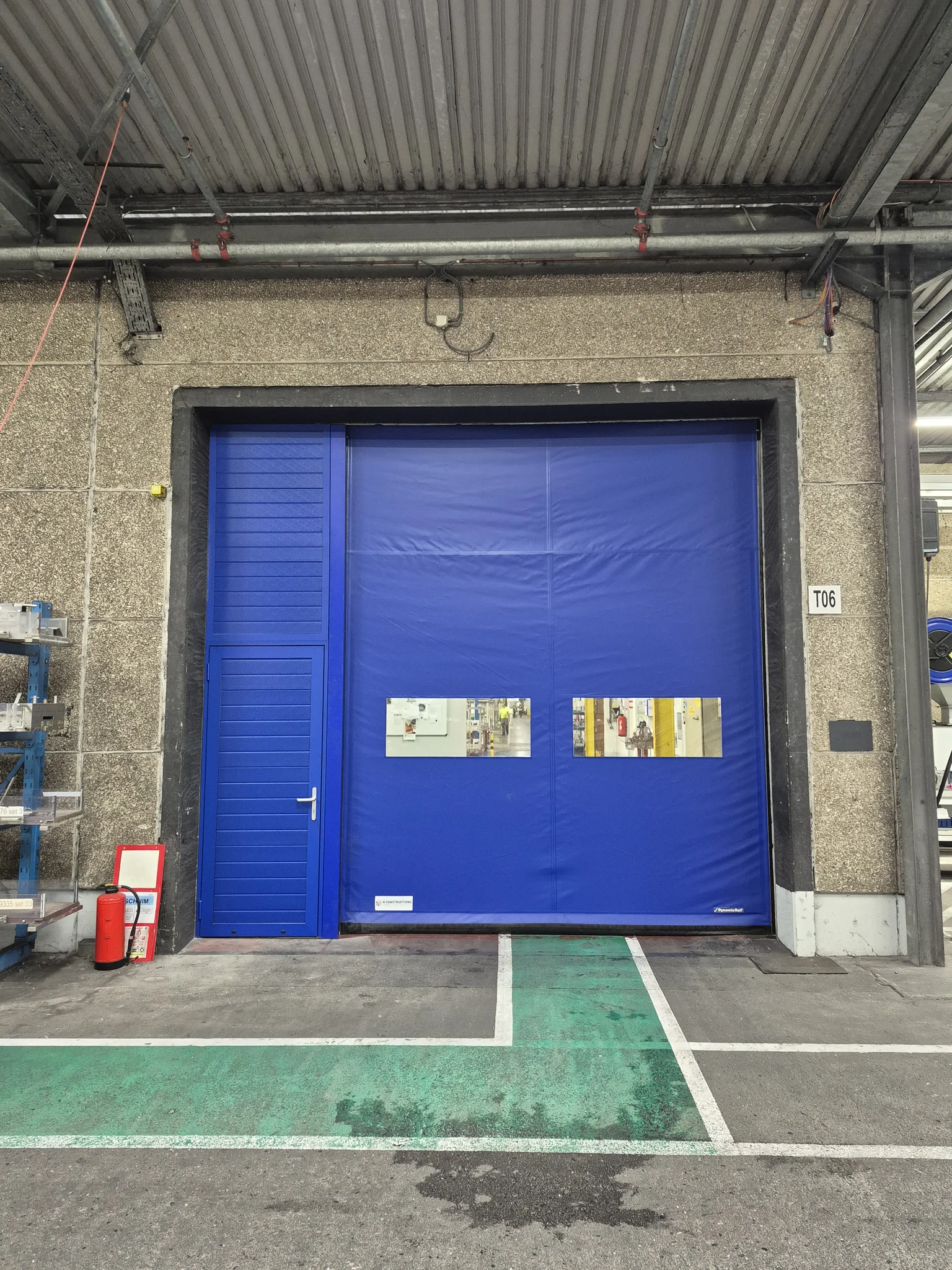 Blauwe industriële garagedeur met zijdeur in een betonnen loods, rode brandblussers links en groene parkeermarkeringen op straat.