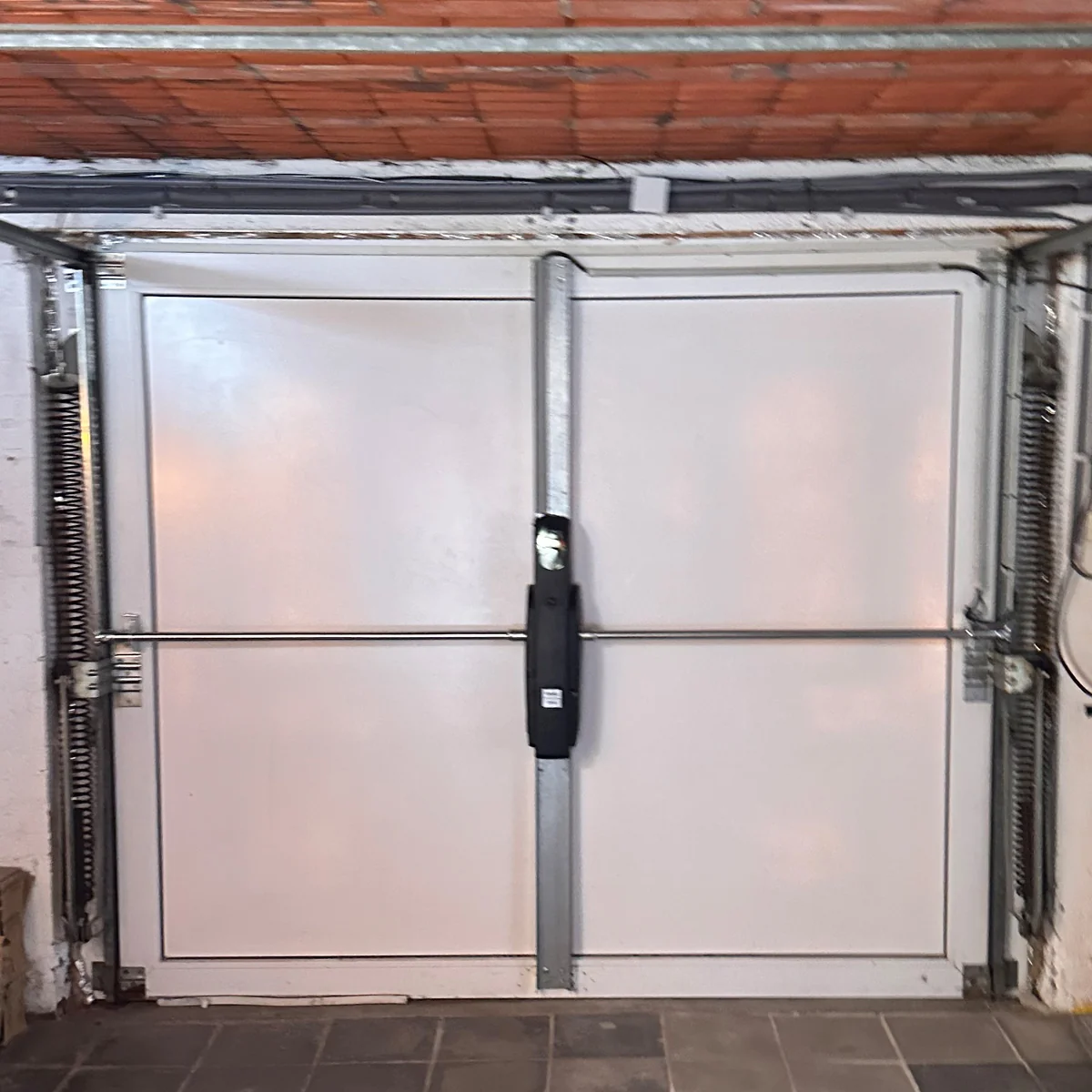 Garagedeur met twee witte panelen en horizontale verdeling, zwarte hefveren op beide zijden en zwarte handgreepstrip in het midden.