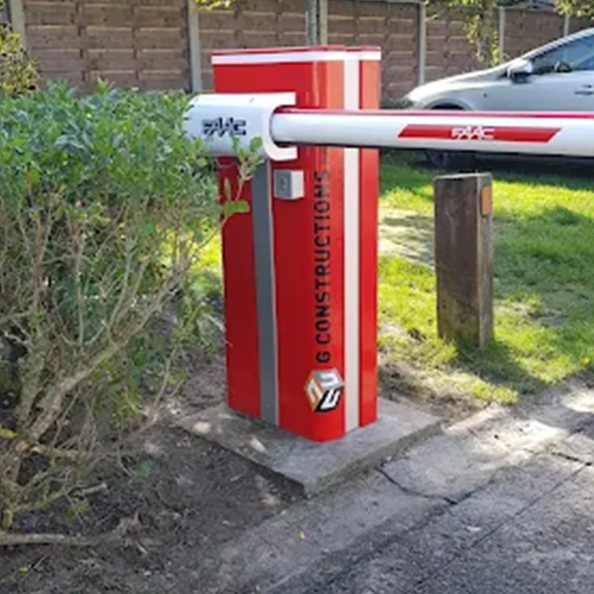 Rode slagboom met wit en rood gestreepte arm bij ingang woonwijk, naast groene haag en parkeerplaats.