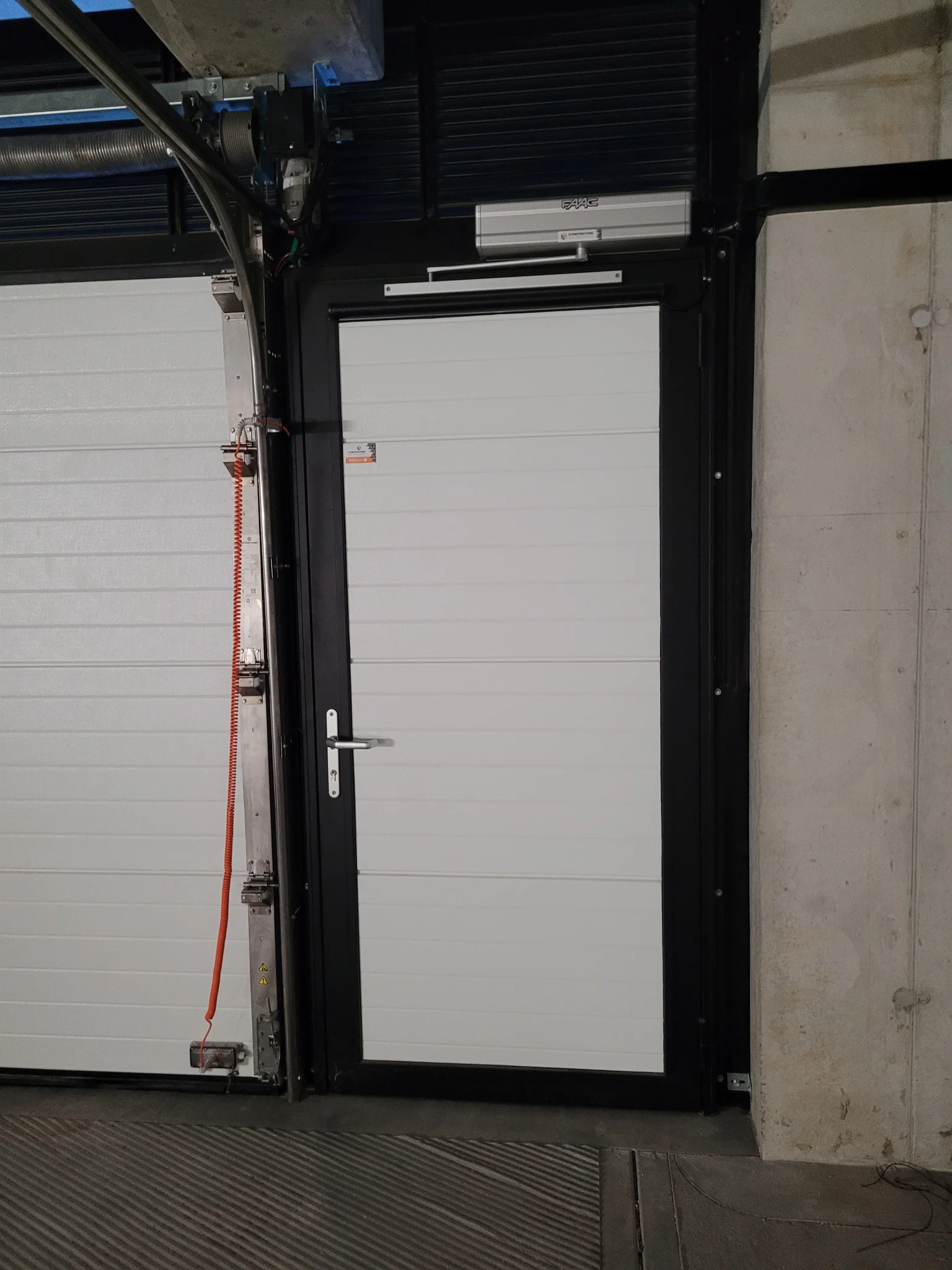 Witte deur met zwart frame en automatische deuropener, geplaatst in betonnen garageopening naast witte garagedeur.
