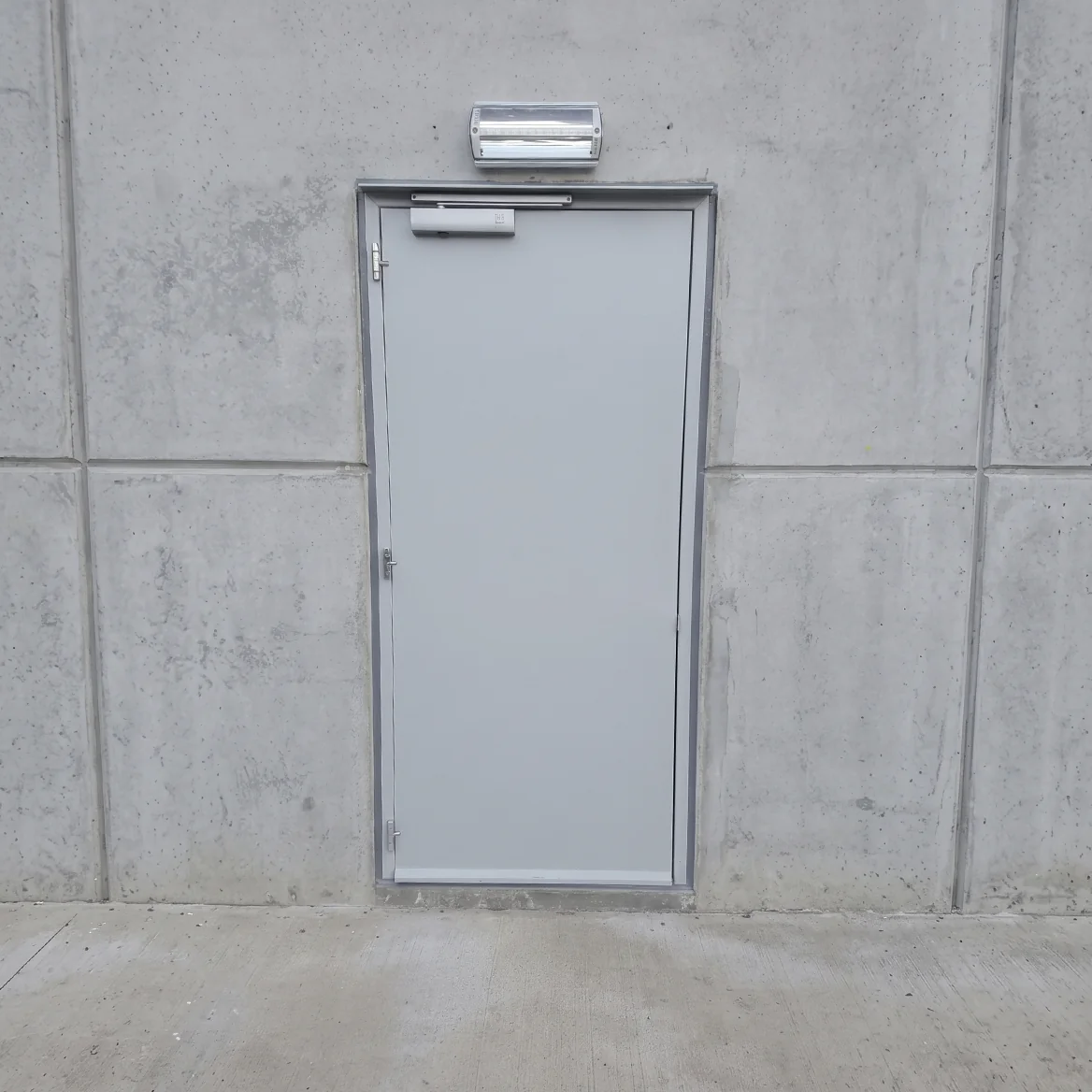 Witte branddeur met grijs frame in betonnen muur, voorzien van drukknop en noodverlichting boven.