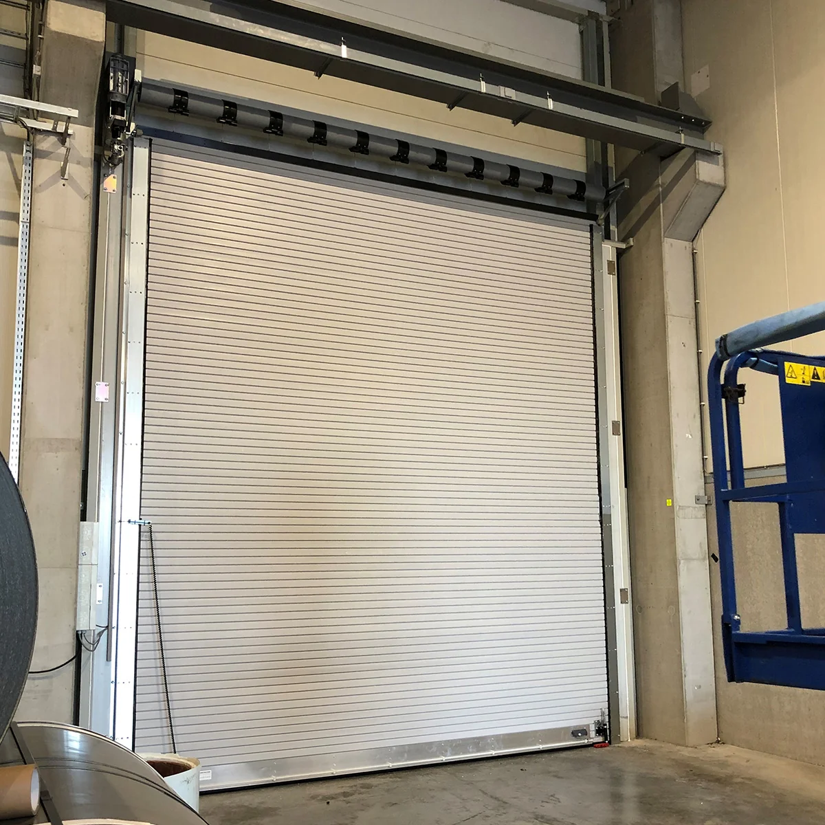 Industriële brandwerende rolluikdeur in wit aluminium met horizontale lamellen in een betonnen deuropening van een bedrijfshal.