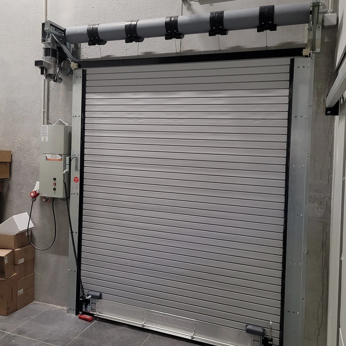 Gesloten grijze brandwerende rolpoort met horizontale lamellen, elektronische besturing aan zijwand en ronde torsie-as bovenaan.