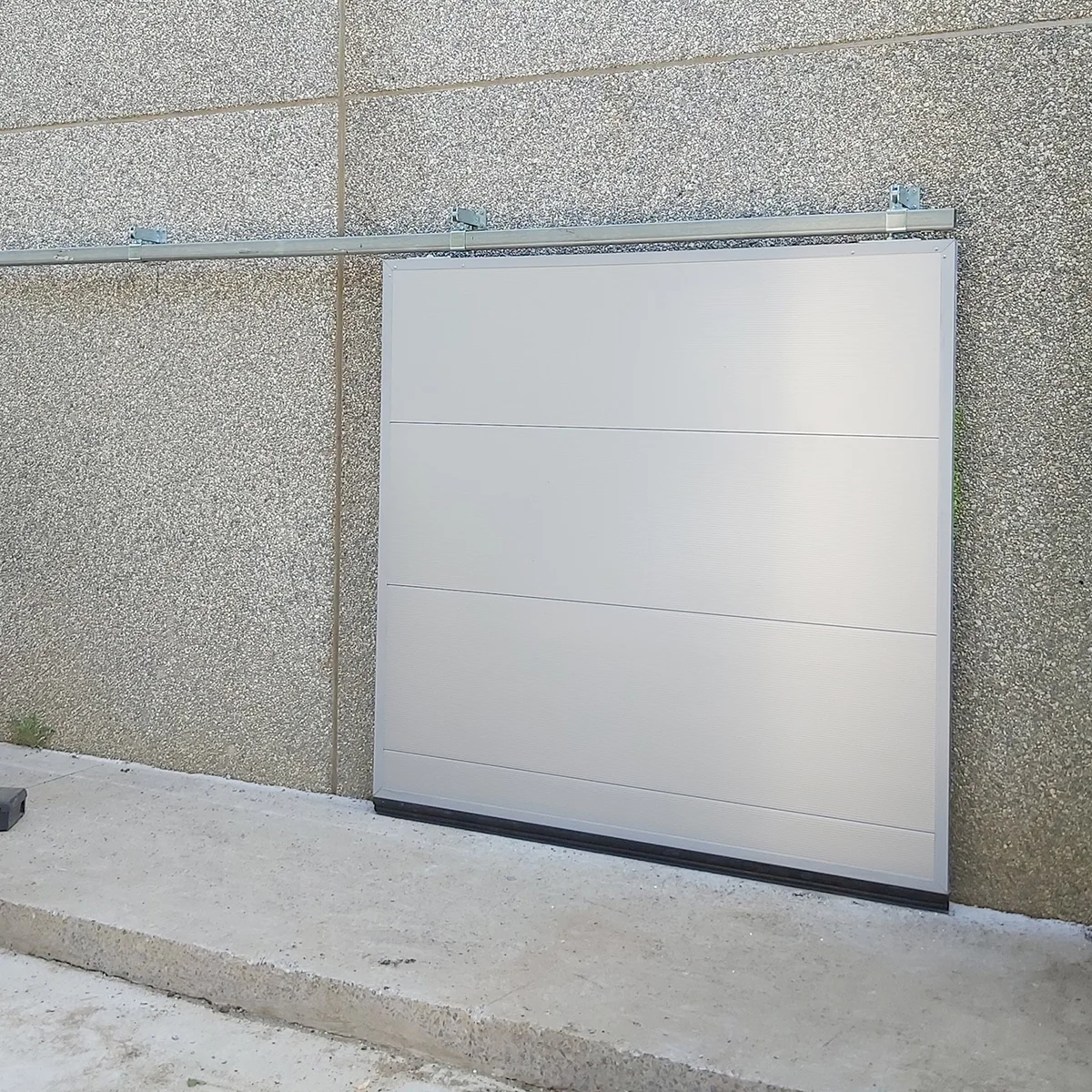 Witte garagedeur met horizontale panelen gemonteerd in een grind- en betonwand met stalen bovenbalk.