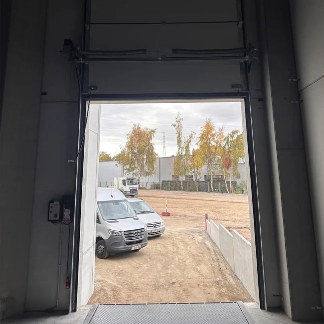 Uitzicht vanuit een open laaddeuren naar een terrein met geparkeerde bestelwagens en berken onder een bewolkte hemel.