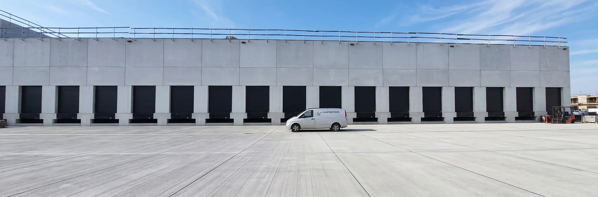 Witte bestelwagen staat voor een groot magazijngebouw met meerdere zwarte laaddeuren en betonnen laadperrons.