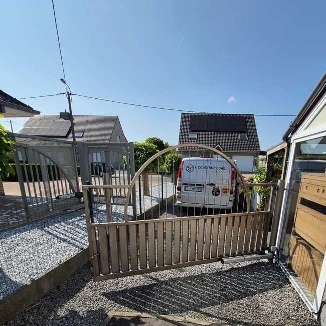 Grijze draaipoort van metaal in een oprit tussen twee huizen met een bedrijfsbus daarachter.