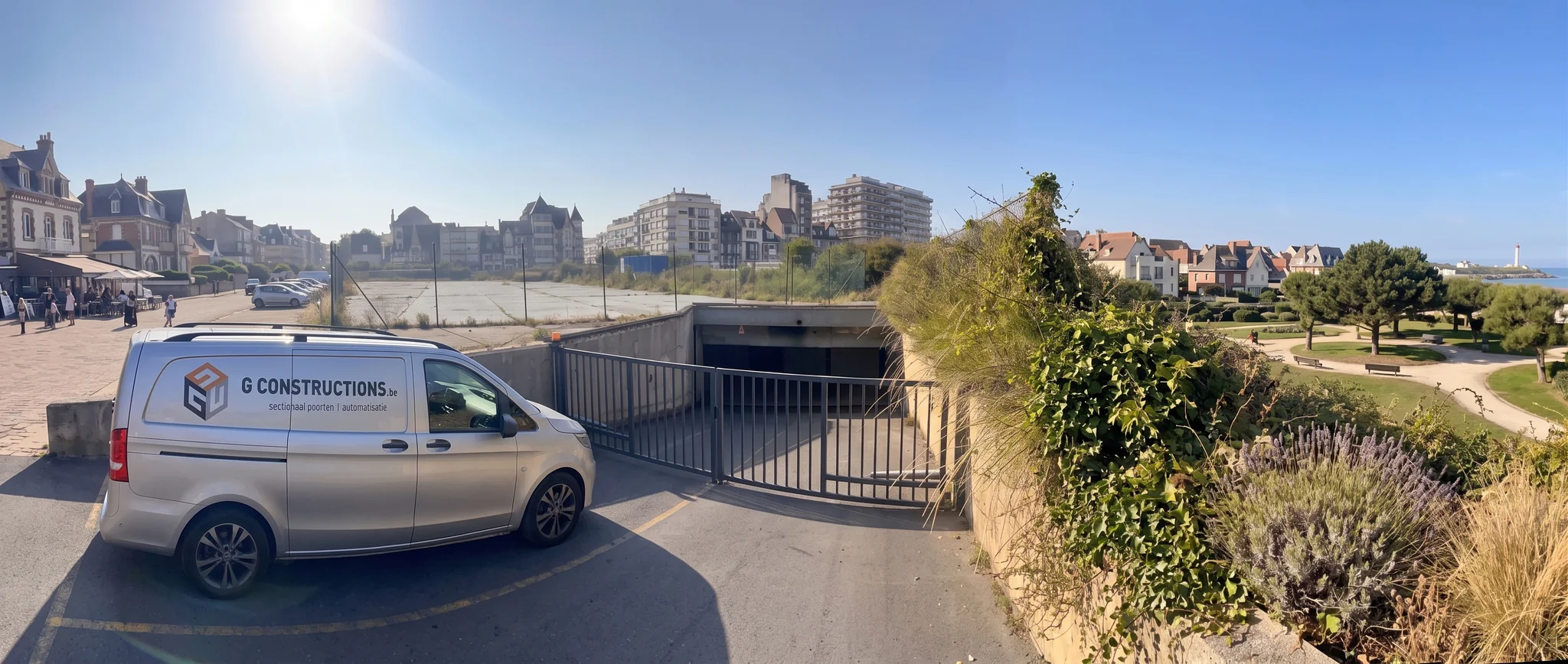 Witte bedrijfsbus van G Constructions staat naast een open draaipoort met zwart metalen hekwerk aan een kustpromenade.