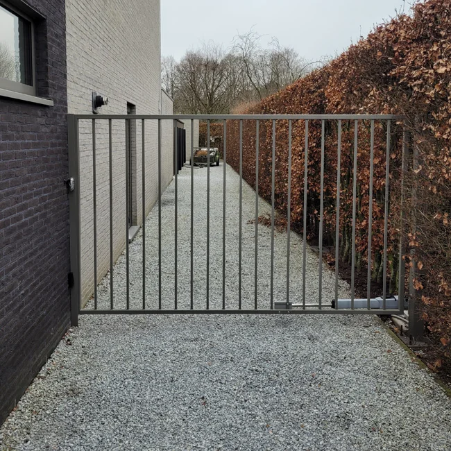 Moderne draaipoort van donkergrijs metaal in een smalle doorgang tussen een bakstenen woning en bruine haag.