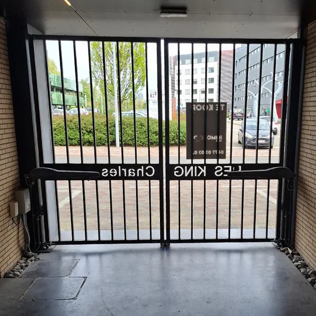 Twee zwarte schuifdeuren met verticale spijlen openen uit op een parkeerplaats met groene bomen en stedelijke gebouwen.