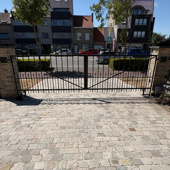 Zwart metalen draaipoort in geopende stand voor parkeerterrein met geparkeerde auto's en woongebouwen op achtergrond.