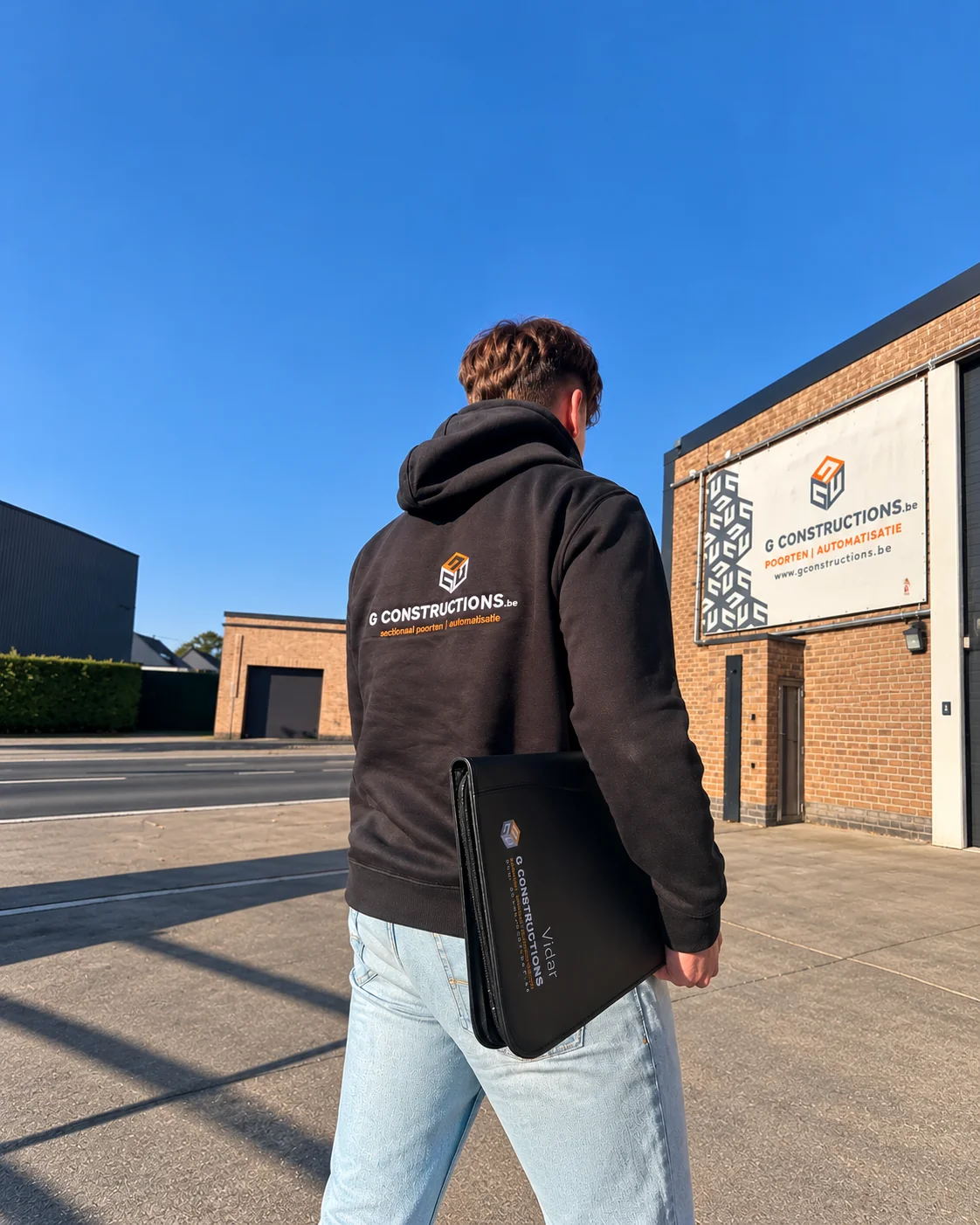 Persoon met rood haar draagt een zwarte hoodie met G Constructions-logo en een zwarte tas, staande op een bedrijfsterrein met bakstenen gebouwen.