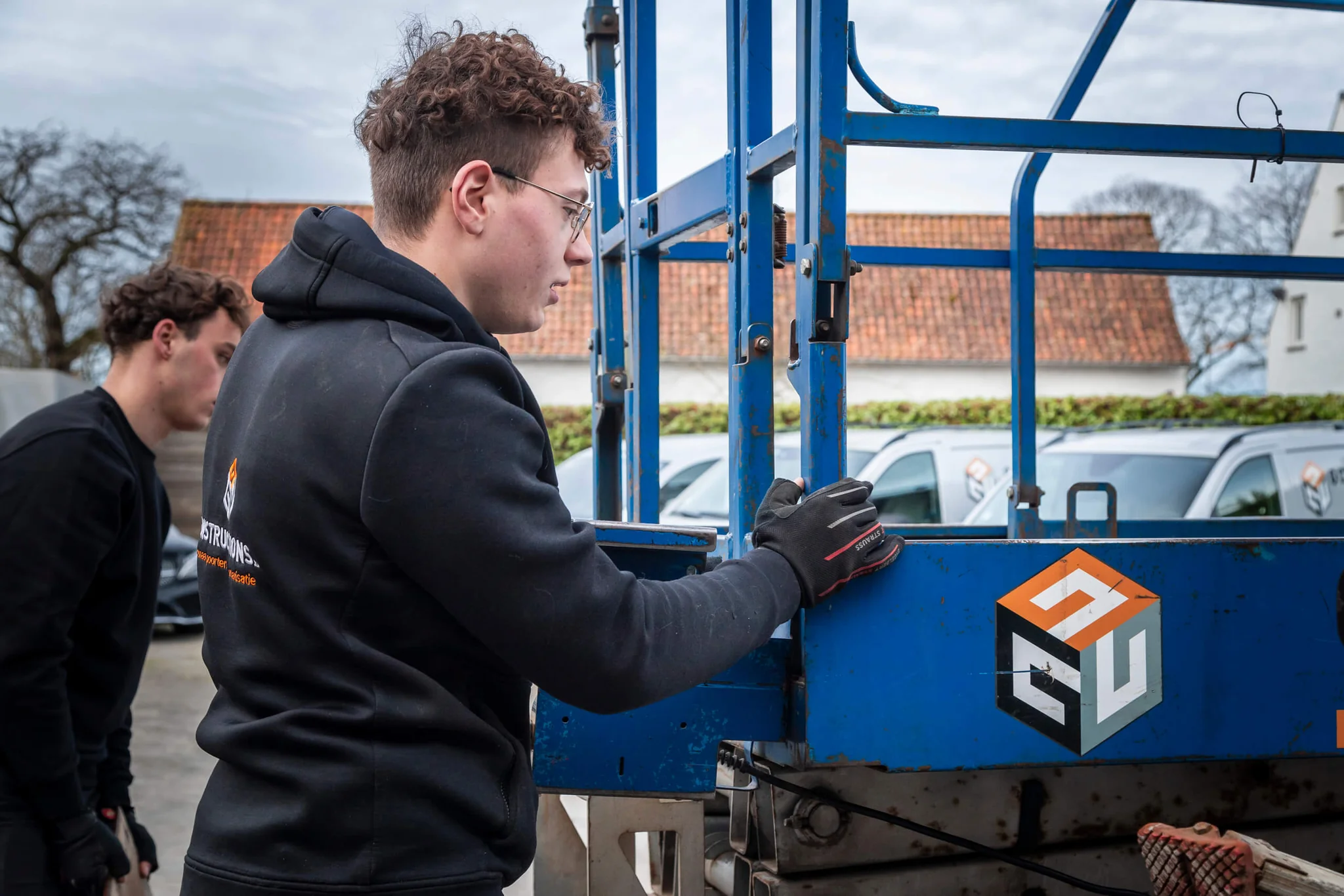 Jongeman in zwarte hoodie en werkhandschoenen beweegt een blauwe aanhanger met oranje logo op een parkeerterrein met huizen op achtergrond.