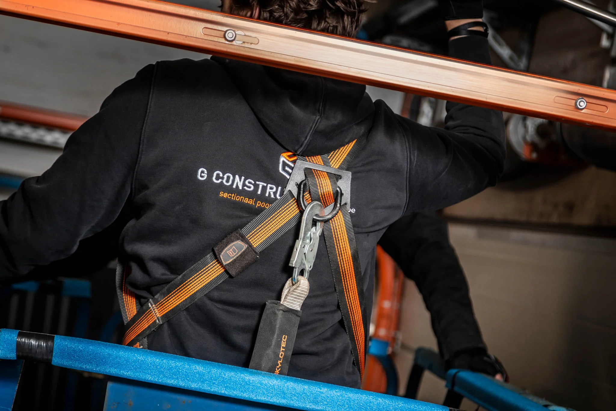 Een zwart veiligheidshesje met oranje strepen en bevestigde veiligheidskabel met karabijnhaak, gedragen door een werker op een bouwplaats.