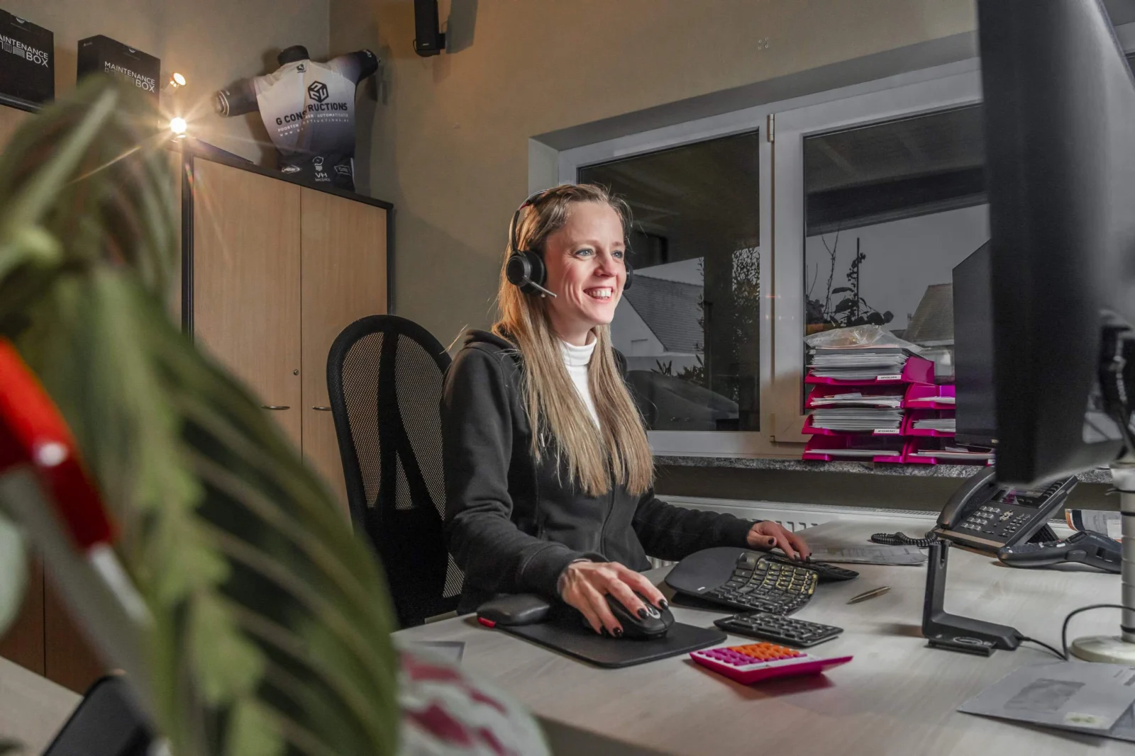 Vrouw met headset zit aan bureau met computer, toetsenbord en muis in een moderne thuiskantoor met plant op voorgrond.