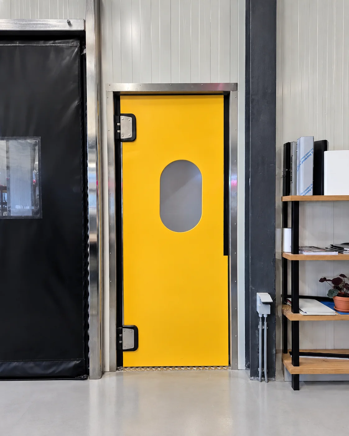 Gele pendeldeur met rond zietraam in industrieel interieur naast zwarte deur en houten stellage.