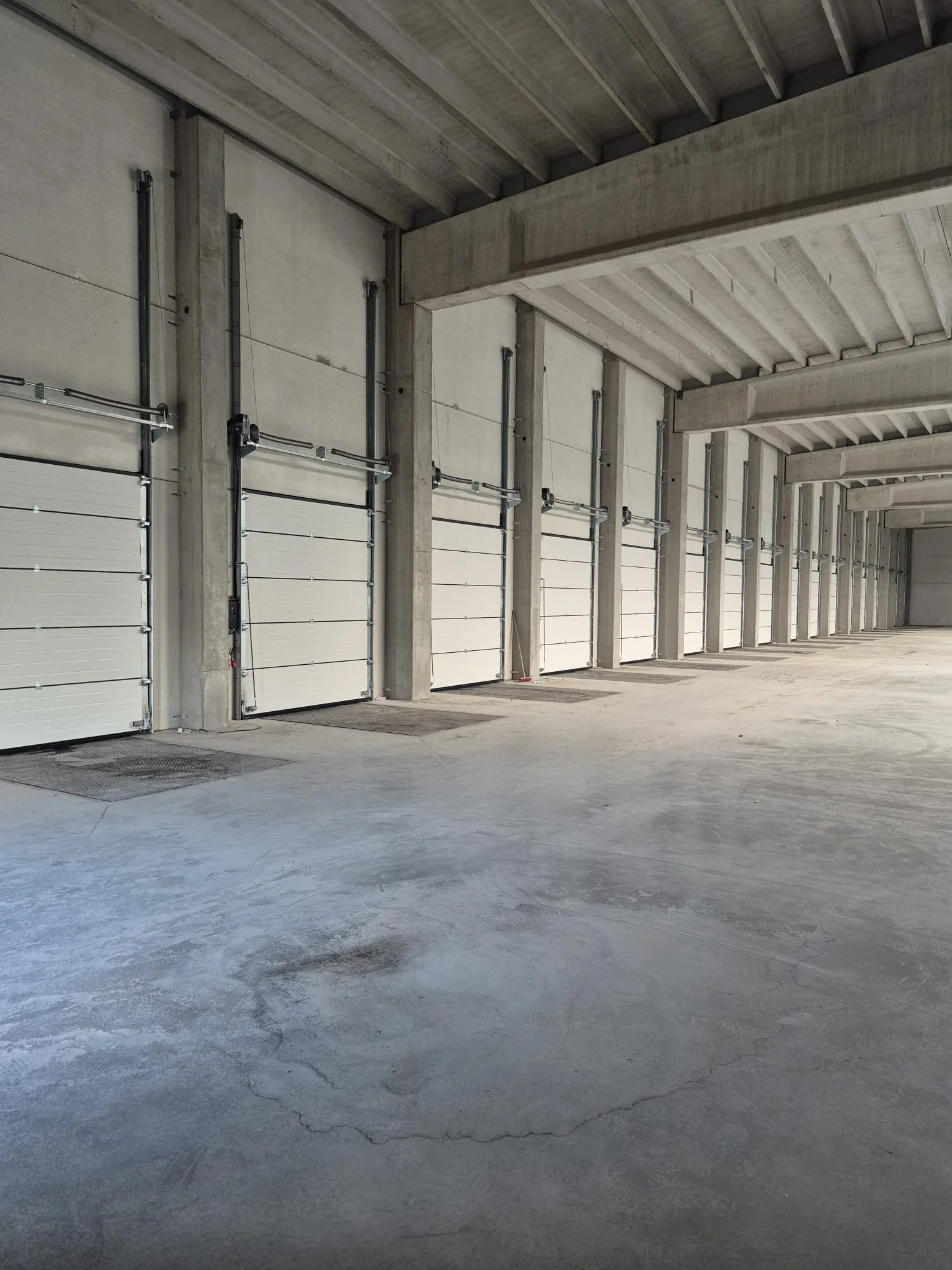Binnenruimte van een industrieel gebouw met betonnen vloer en meerdere witte garagdeuren langs betonnen kolommen.