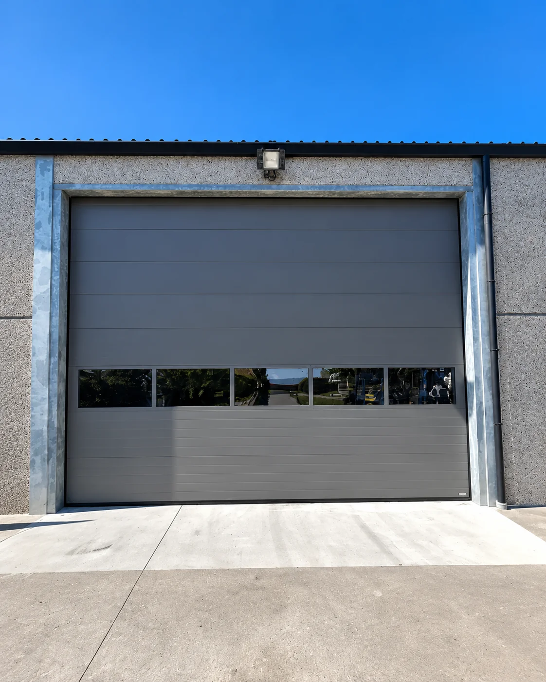 Grijze garagedeur met glaspanelen in een betonnen muur onder blauwe hemel.