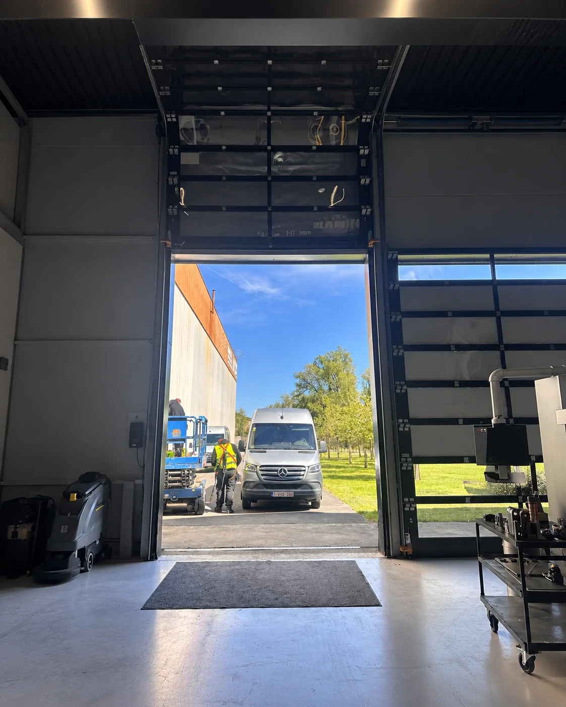 Zicht vanuit een werkplaats door open deuren naar buiten met een Mercedes-bestelwagen en werknemer in geel hesje op het terrein.