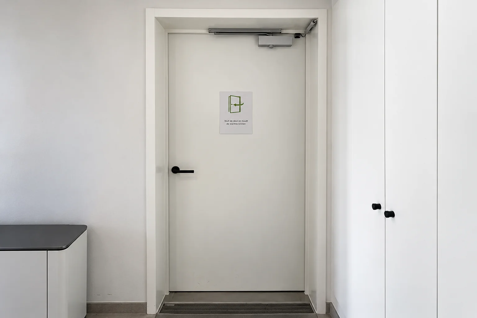 Witte branddeur met zwart handvat en automatische deursluiters bovenaan, waarop een instructiebord is bevestigd.