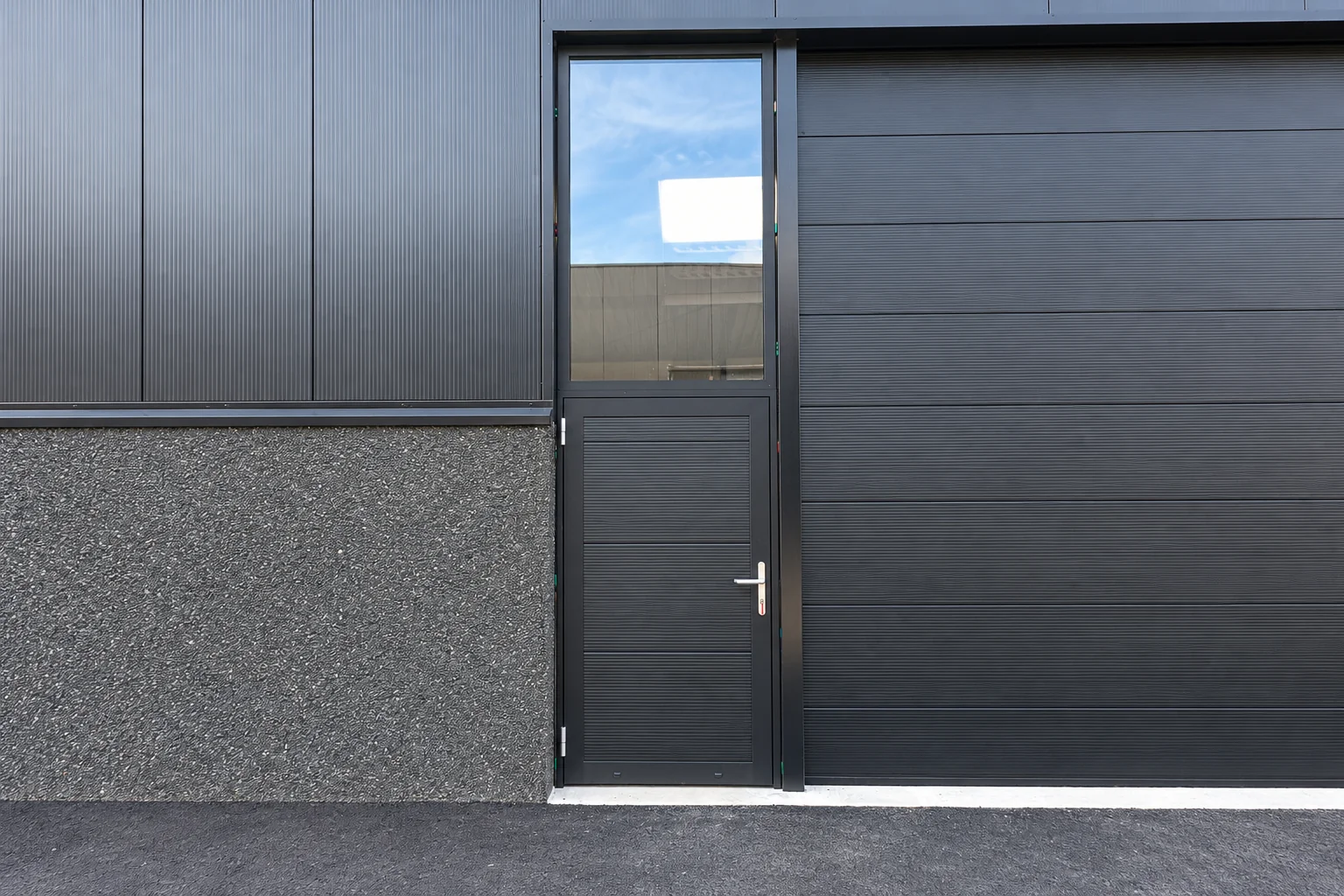 Moderne grijze garagedeur met daarnaast een zwarte aluminium loopdeuren met raam en horizontale lamellen.
