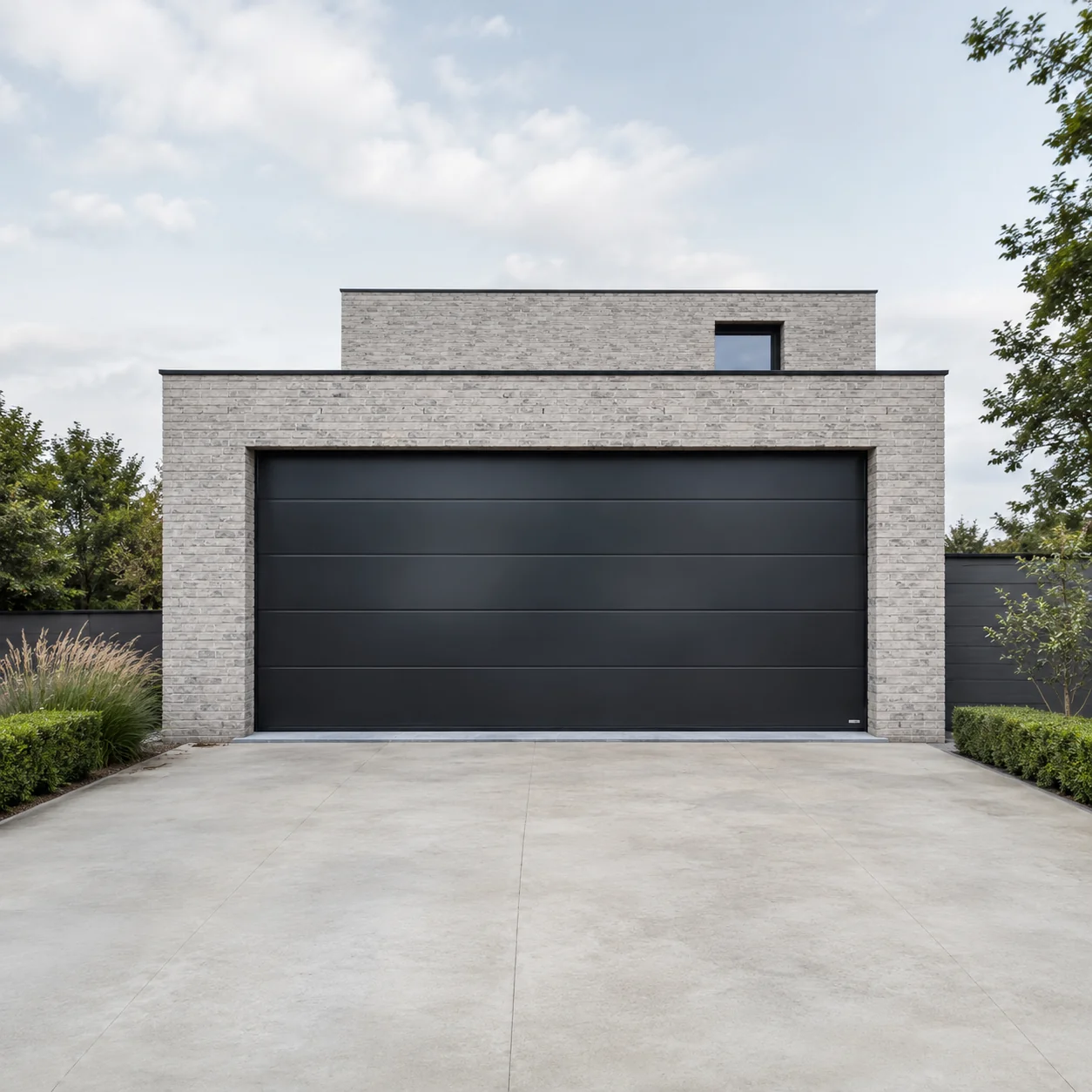 Modern huis met grijze baksteengevel en grote zwarte sectionaalpoort op betonnen oprit omgeven door groene struiken.