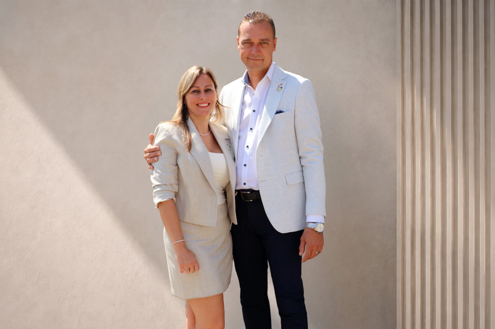 Een man in lichtblauw blazer met donkerblauwe broek en een vrouw in beige blazer staan glimlachend naast elkaar tegen een witte muur.