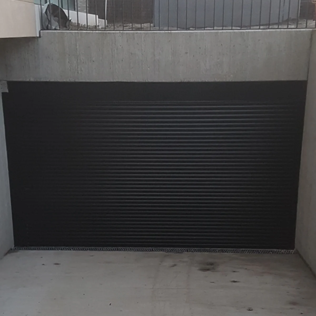 Zwarte roldeur met horizontale lamellen in een betonnen deuropening met hekwerk erboven.
