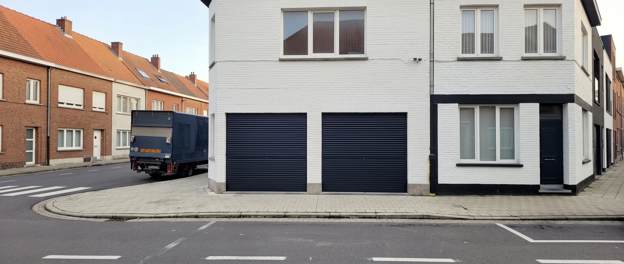 Witte woning met twee donkerblauwe rolpoorten voor garageboxen aan straatgevel naast rood bakstenen huizen.