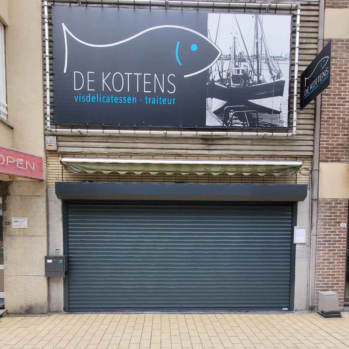 Gesloten metalen roldeur van een restaurant genaamd De Kottens met visdelicatessen en traiteur, met een vintage scheepsboot op het uithangbord.