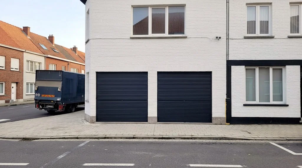 Witte woning met twee zwarte rolpoorten in de garageopening aan de straatzijde, naast bakstenen huizen en een bestelwagen.