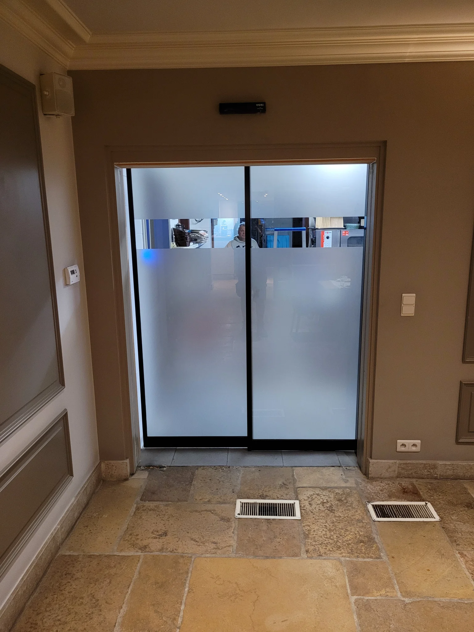 Automatische schuifdeuren met mat glas en zwart frame in een beige hal met stenen vloer.