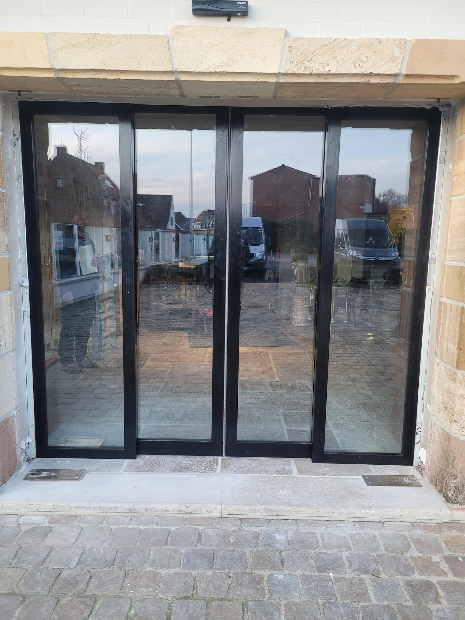 Vier zwartgrijze schuifdeuren met glas geven uitzicht op een patio met bakstenen gebouwen en voertuigen.
