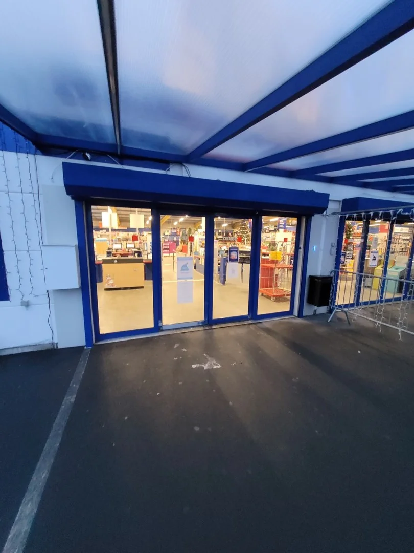 Ingang van een Hubo bouwmarkt met blauwe afdak, glazen deuren en verlichte winkelruimte aan het einde van de dag.