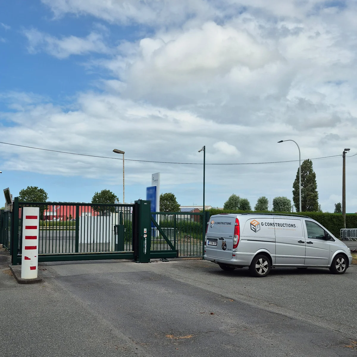 Witte bestelwagen van G Constructions staat voor een groen metalen schuifhek met rood-witte markering op een bedrijventerrein.