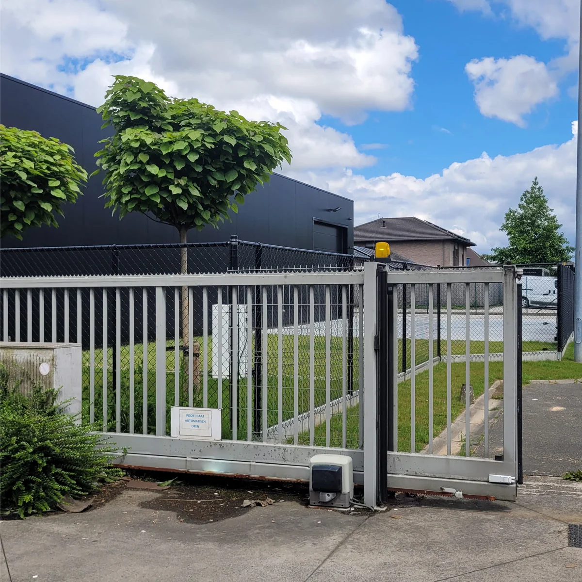 Grijze en witte schuifpoort met roosterwerk bij industrieel terrein, achter moderne gebouwen met groene bomen.