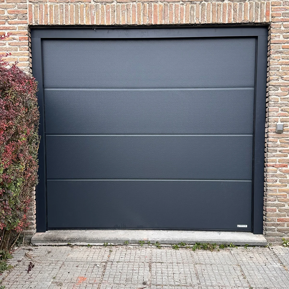 Moderne zwarte sectionaaldeur met horizontale paneelindeling in bakstenen gevel, links bekleed met klimop.