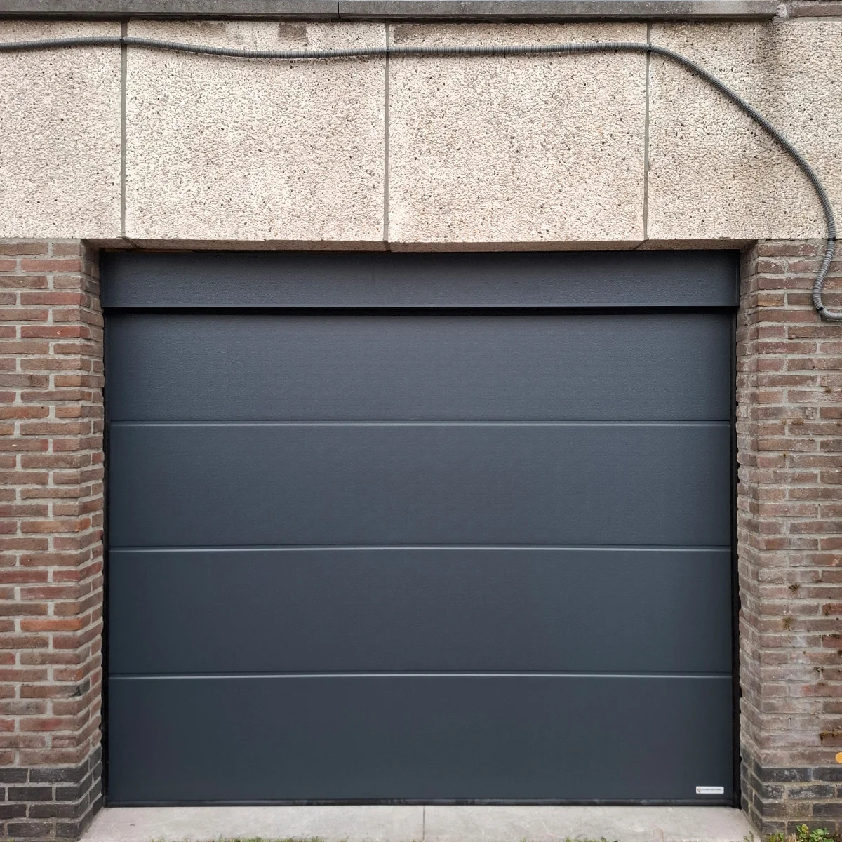 Zwarte sectionaaldeur voor garage met horizontale lamellen, ingebouwd tussen rode bakstenen zijmuren en beige stenen bovenwand.
