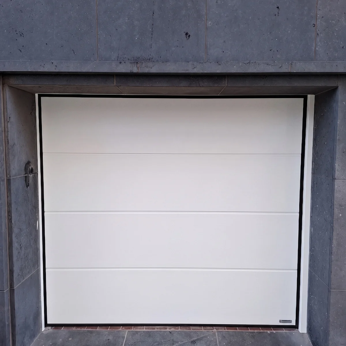 Witte sectionaaldeur in grijze betonnen garageopening met horizontale paneelindeling en zwarte frame.
