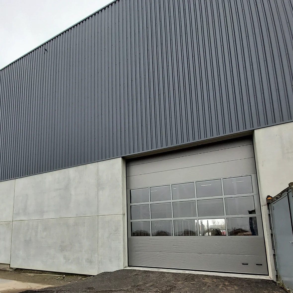 Moderne industriële hal met grijze sectionaaldeur met glaspanelen en golfplaten dak boven betonnen wand.