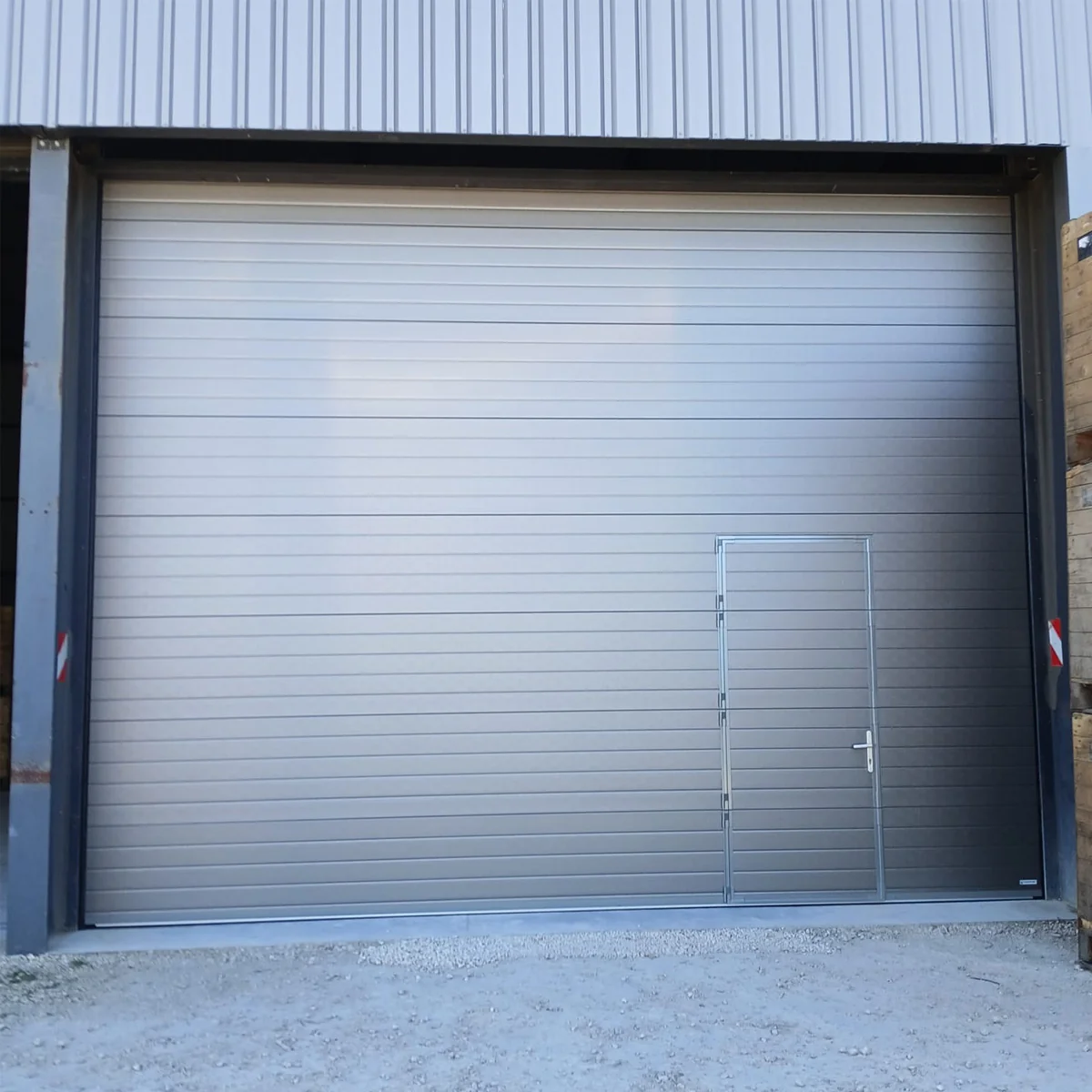Grote industriële sectionaaldeur in grijs aluminium met horizontale lamellen en ingebouwde toegangsdeur op een betonnen ondergrond.