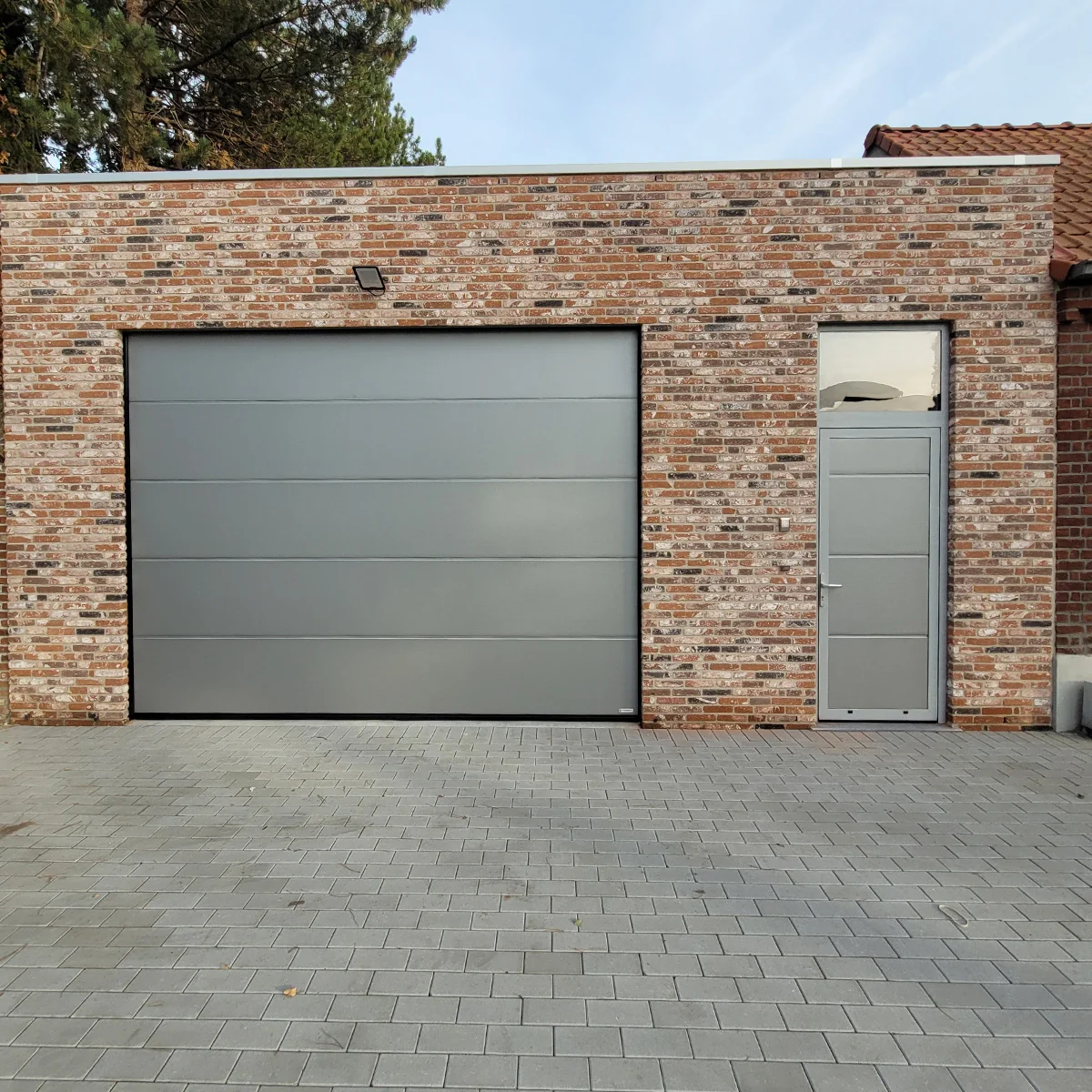 Moderne sectionaaldeur in grijs op bakstenen garagebouw met aangrenzende deur en grijze tegelverharding.