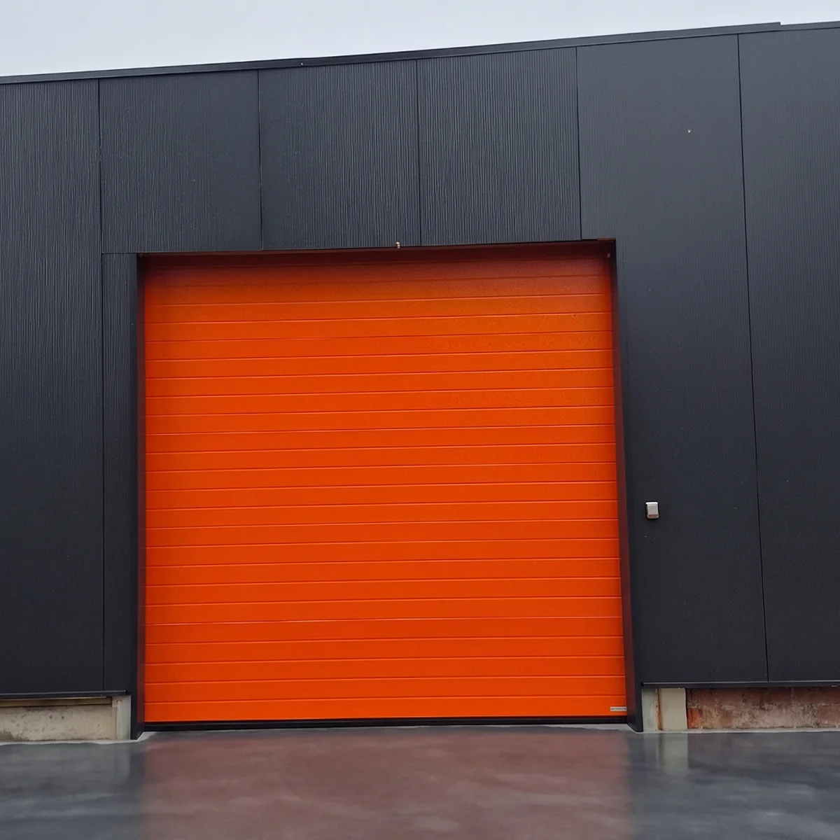 Industriële sectionaaldeur in oranje kleur geïntegreerd in een donkergrijze metalen gevel van een gebouw.