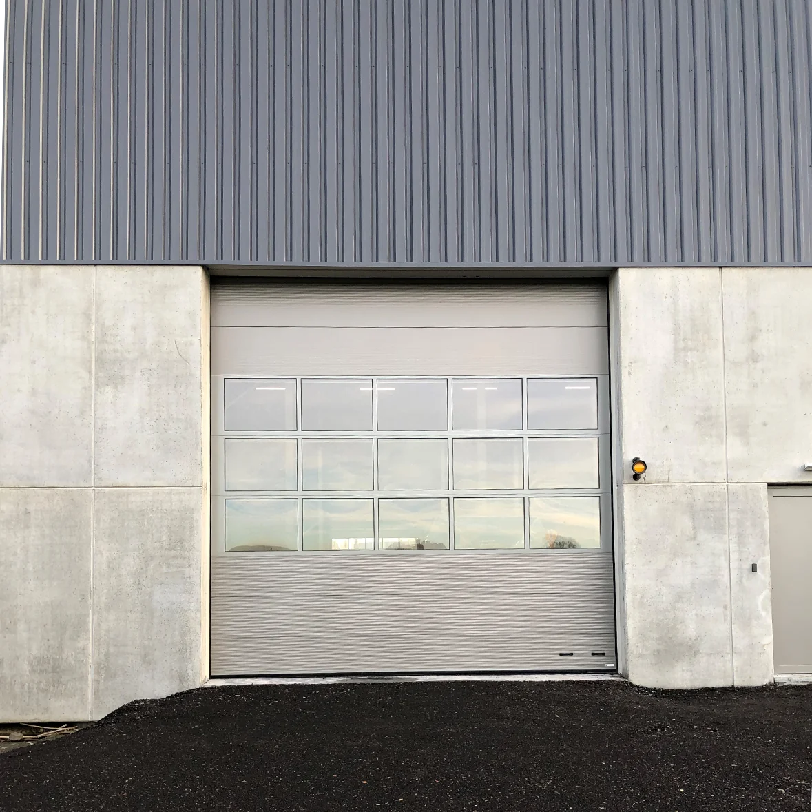 Grijze sectionaaldeur met ramen op industrieel betonnen gebouw met golfplaten dak en donker asfalt.