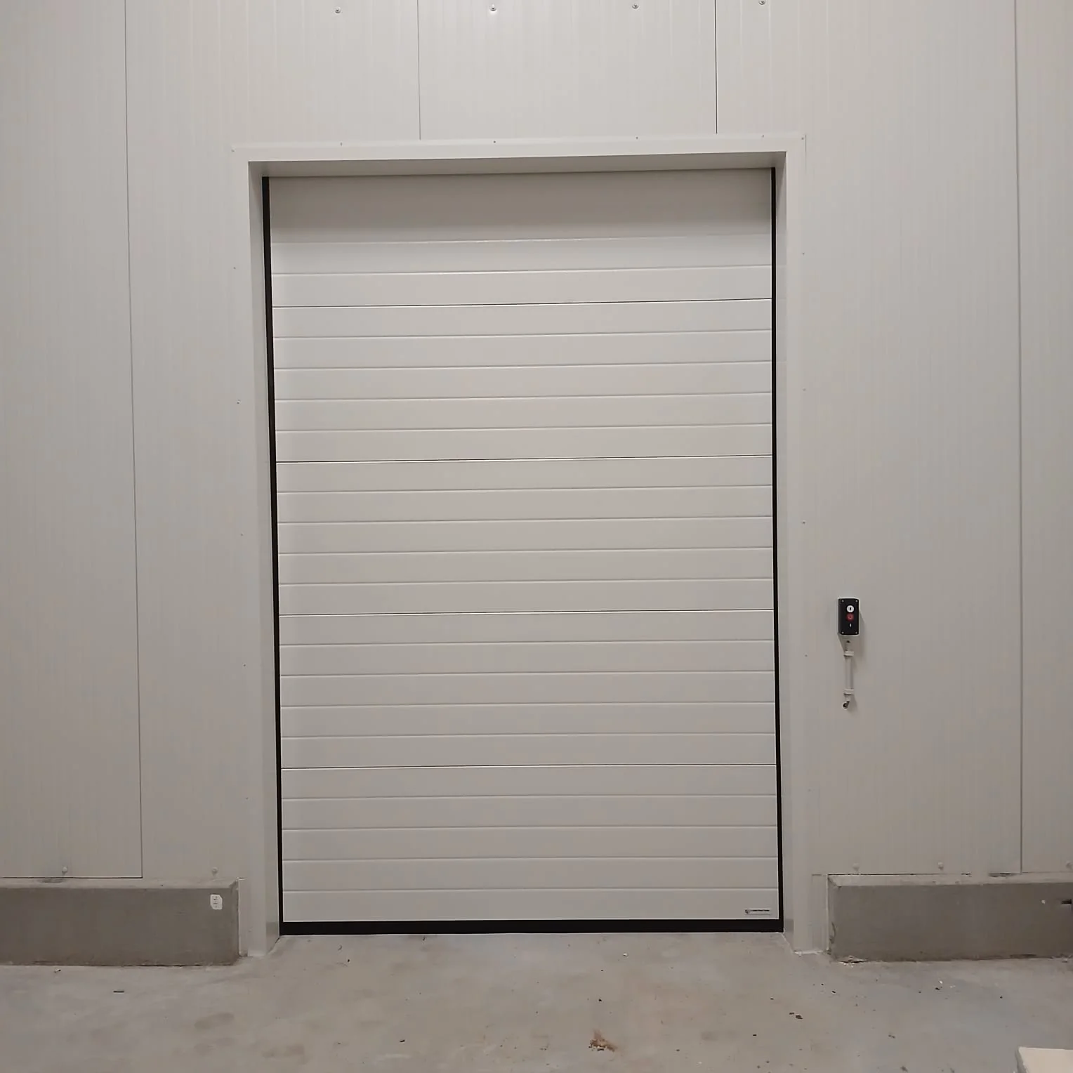 Witte sectionaalpoort met horizontale panelen en zwarte onderrand in betonnen garageopening met bedieningspaneel rechts.