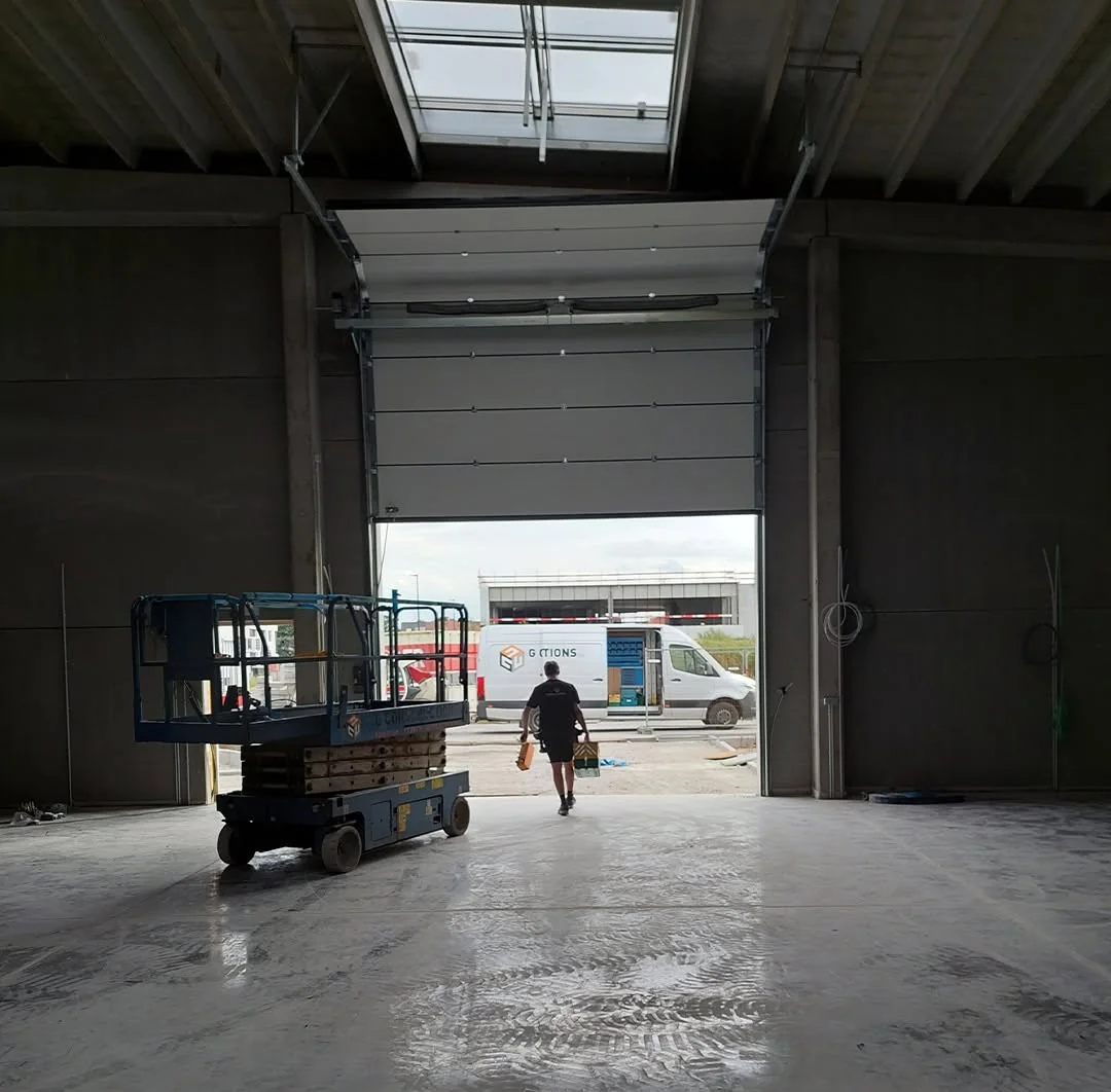 Werkplaats met open garagedeur, een persoon en een blauwe hefplatform op natte betonvloer, bouwplek zichtbaar buiten.