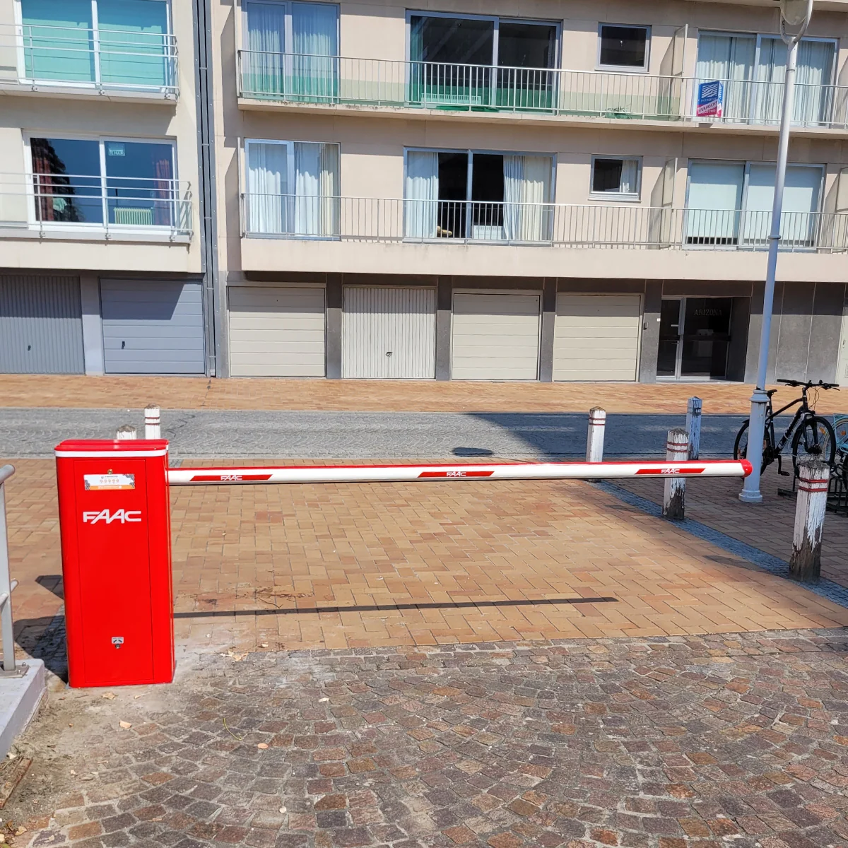 Rode FAAC-slagboom met wit-rood gestreepte arm blokkeert de ingang naar een parkeerplaats voor een modern appartementengebouw.